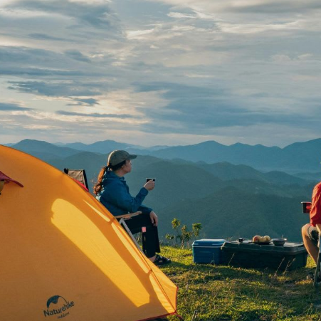 An orange Nemo tent is pitched on a mountain ridge with two people sitting in camping chairs nearby, overlooking layered blue mountain ranges under a cloudy sky.