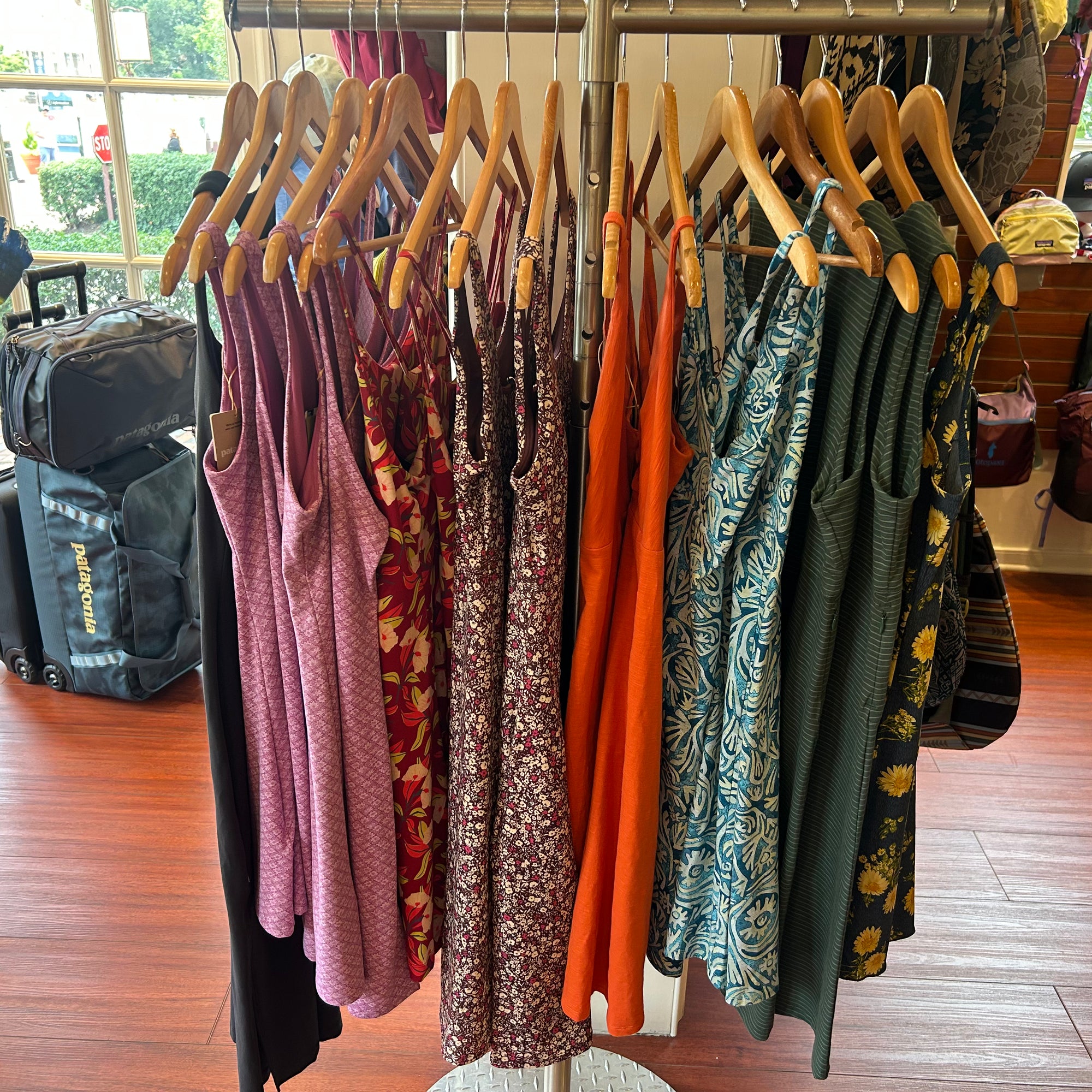 A clothing rack in a retail store displaying a variety of sleeveless dresses in different colors and patterns, including pink, floral, orange, and green, hanging on wooden hangers over a polished wood floor.